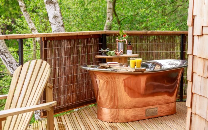 Outdoor copper bath on a treehouse terrace at Treetop Hideaways, with a wooden Adirondack chair and a drinks tray set across the bath, screened by birch woodland